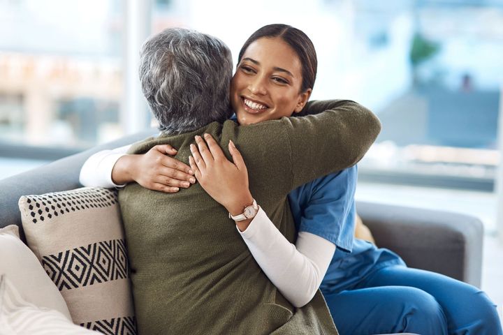 A smiling caregiver hugs an elderly person on a couch. They are in a brightly lit room, likely at home.