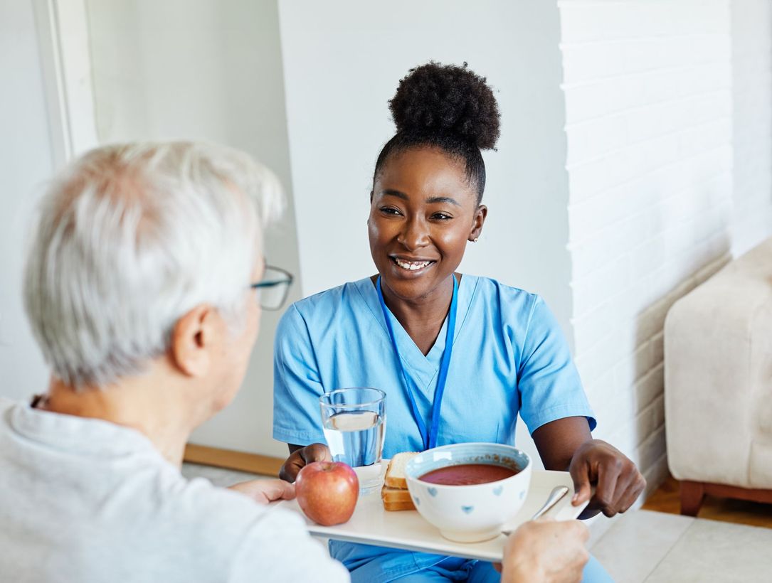 An elderly man and a smiling nurse look at a tablet while seated on a sofa. The nurse points at the tablet.