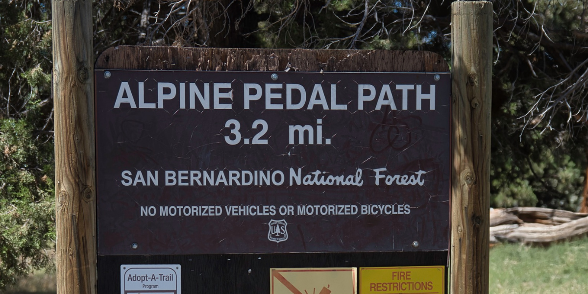 Weathered sign reading “Alpine Pedal Path 3.2 mi” in San Bernardino National Forest, among trees