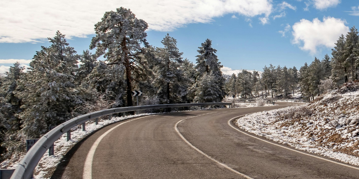 Winding road through snow-covered trees under a blue sky with clouds.