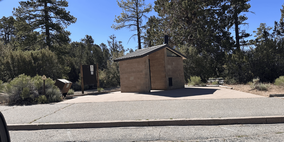 Small tan building with a slanted roof at Woodland Trail, surrounded by trees and shrubs under a blue sky