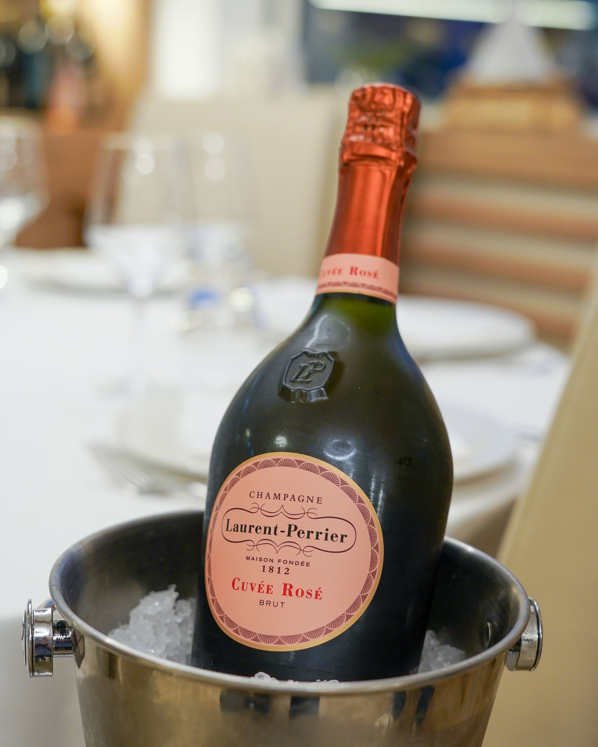 Bottiglia di champagne in un secchiello del ghiaccio su un tavolo, con collo in lamina rossa ed etichetta rosa.