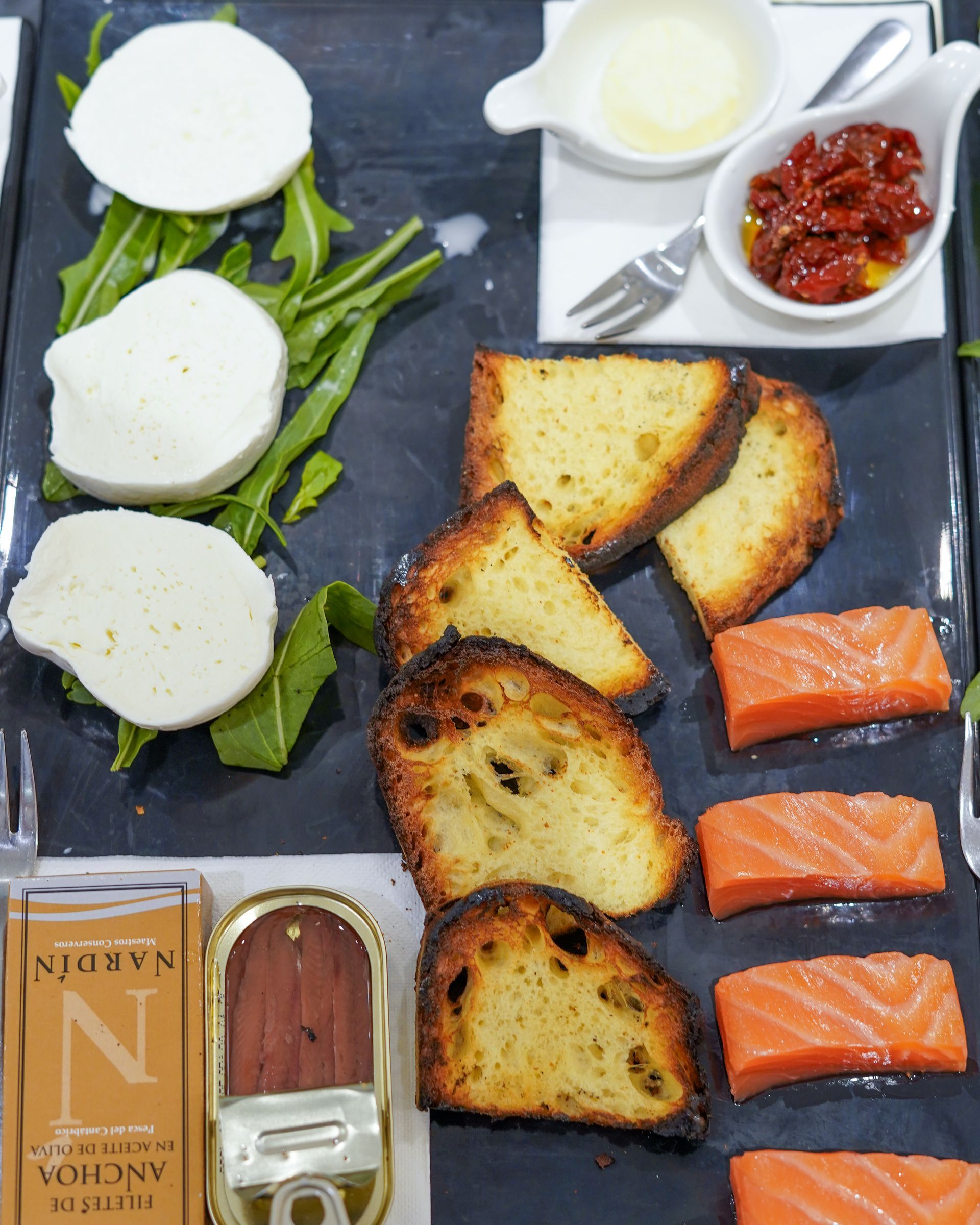 Piatto di sushi assortito con salmone, tortini di pesce, pane, verdure, salsa e burro su un vassoio scuro.