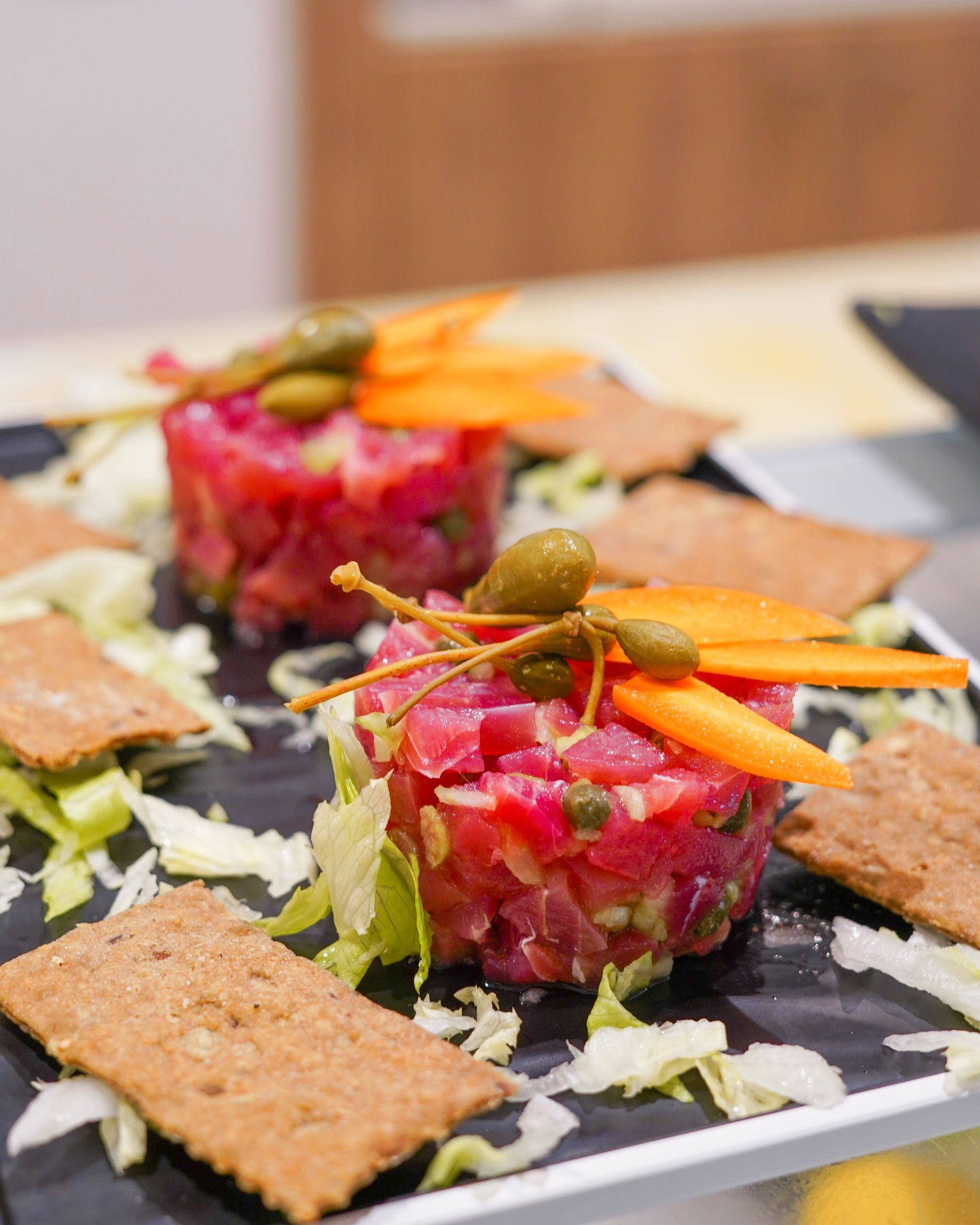 Tartare di tonno gourmet servita su un piatto di ardesia nera con cracker, lattuga tritata e striscioline di carota.