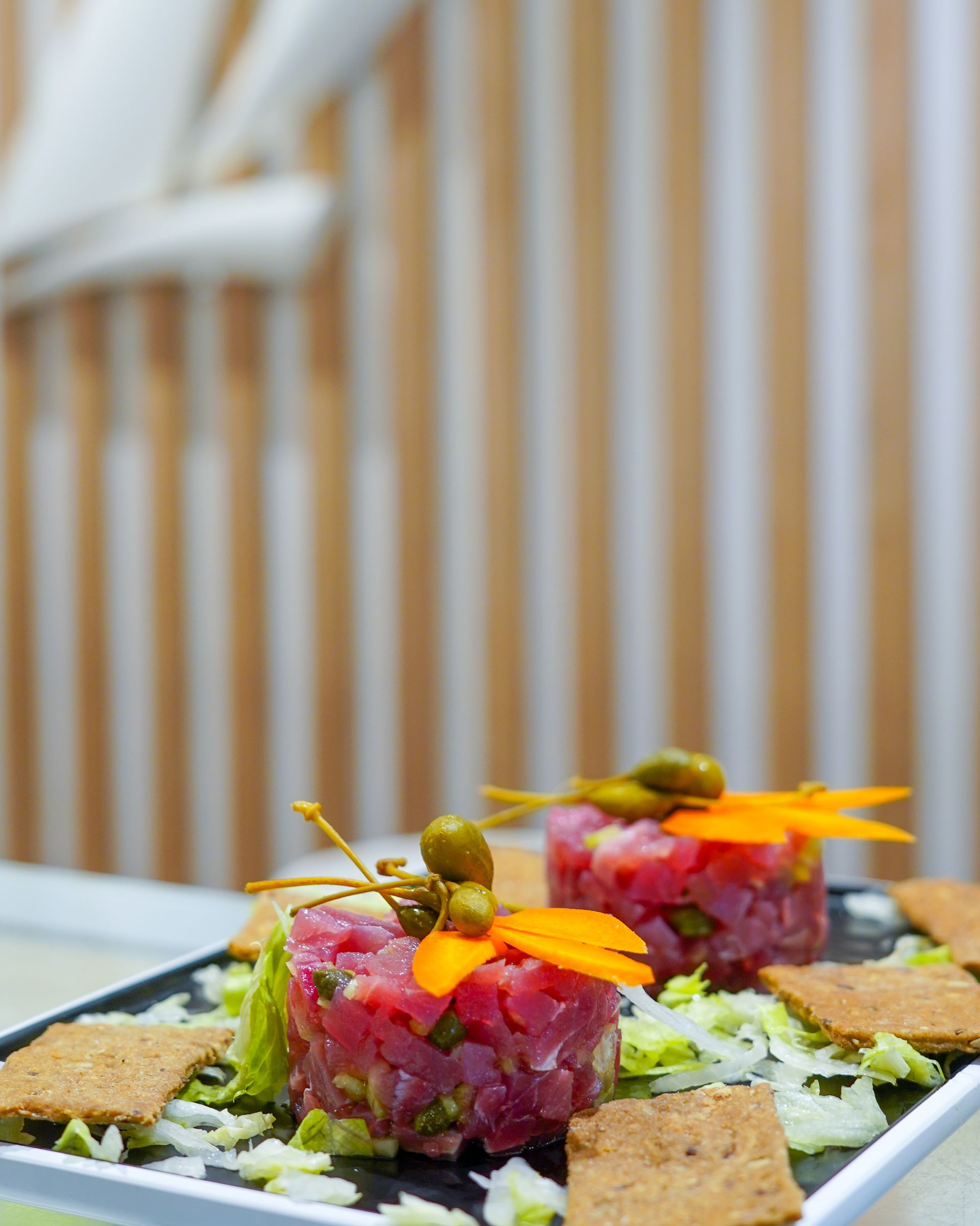 Tartare gourmet con fiori edibili su un piatto bianco, servita su uno sfondo di legno sfocato.