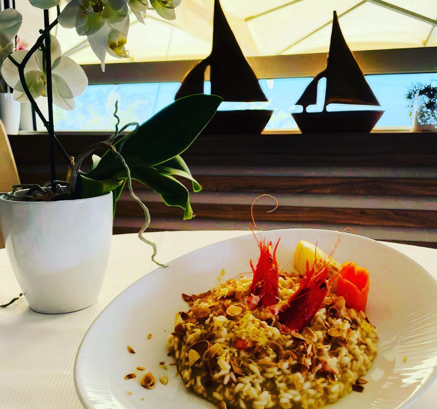 Un piatto di risotto con gamberi rossi su un tavolo accanto a un'orchidea bianca e a una decorazione a forma di barca a vela, vicino a una finestra.