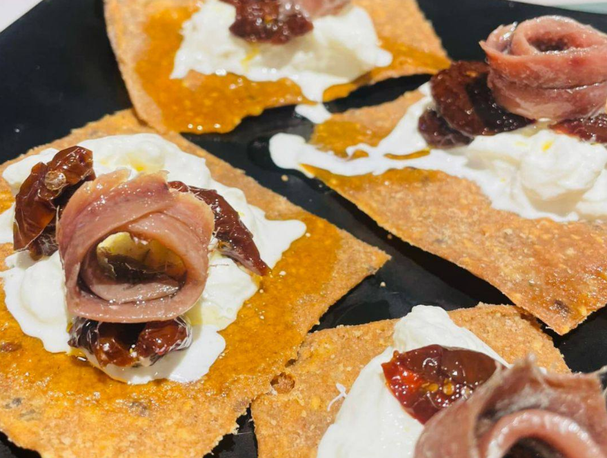 Crackers croccanti all'arancia ricoperti di formaggio bianco e guarniti con acciughe arrotolate, serviti su un piatto scuro.