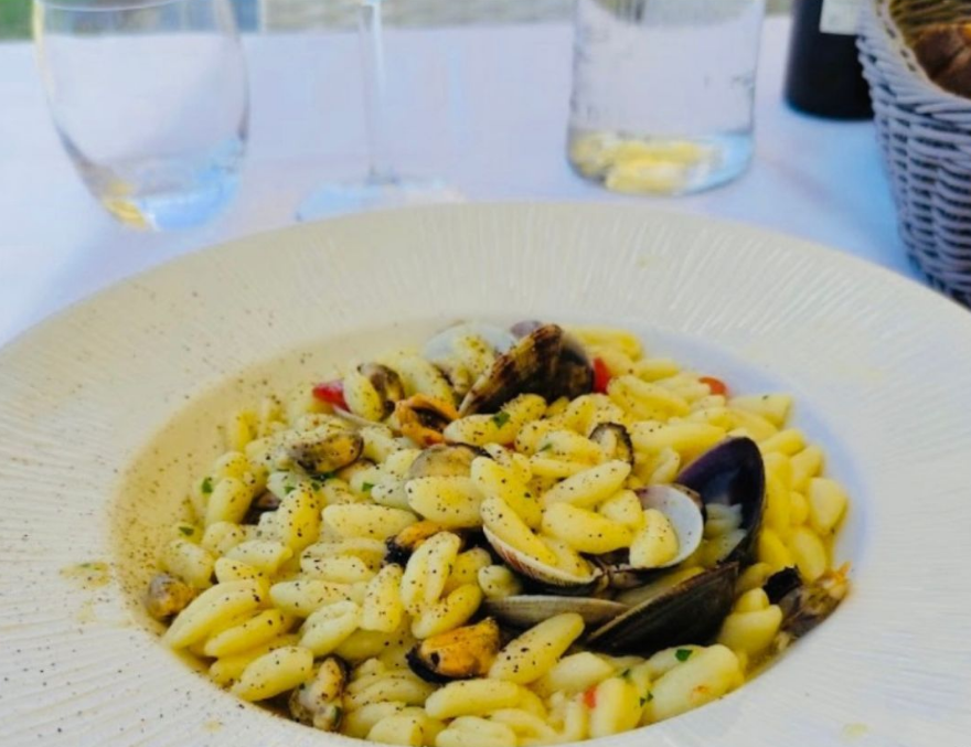 Un piatto di pasta con cozze ed erbe aromatiche su un tavolo, con dei bicchieri sullo sfondo.