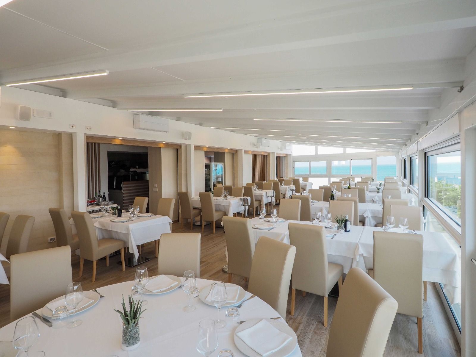 Elegante sala da pranzo di un ristorante sul mare, con tovaglie bianche, sedie color crema e ampie finestre che si affacciano sull'acqua.