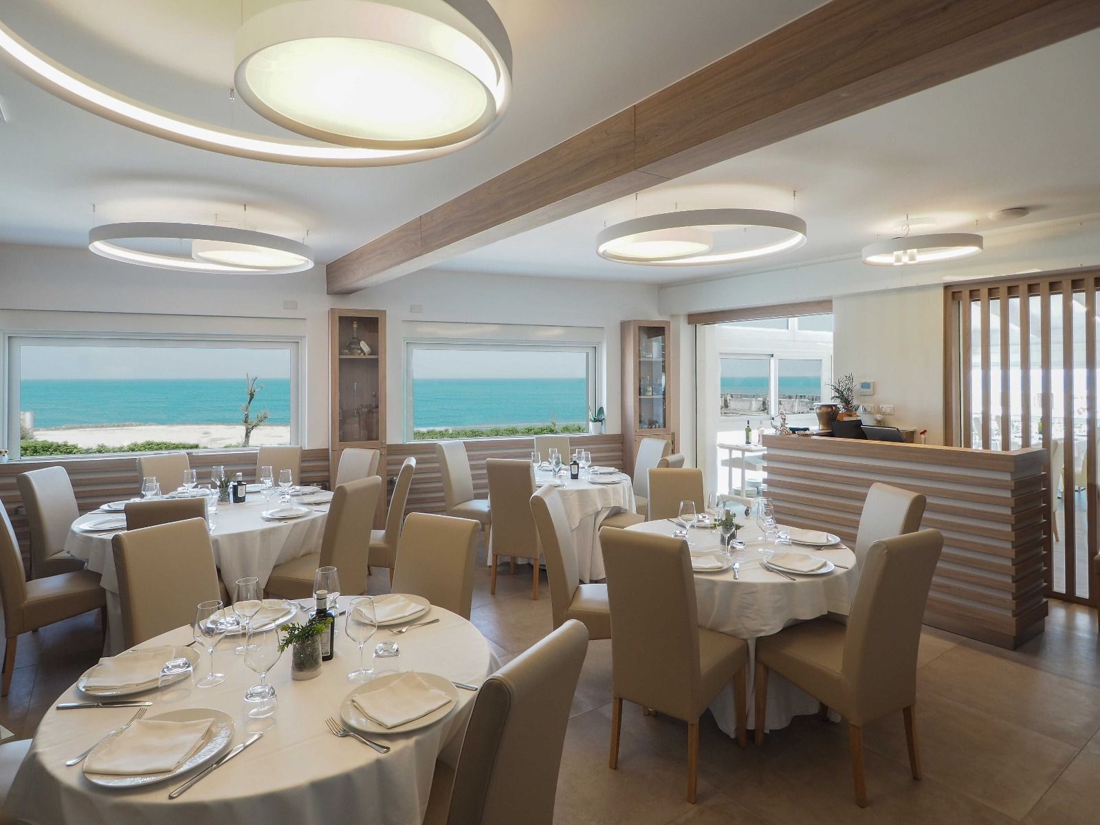 Elegante sala da pranzo di un ristorante sul mare, con tavoli apparecchiati, sedie color crema e finestre con vista sull'oceano.