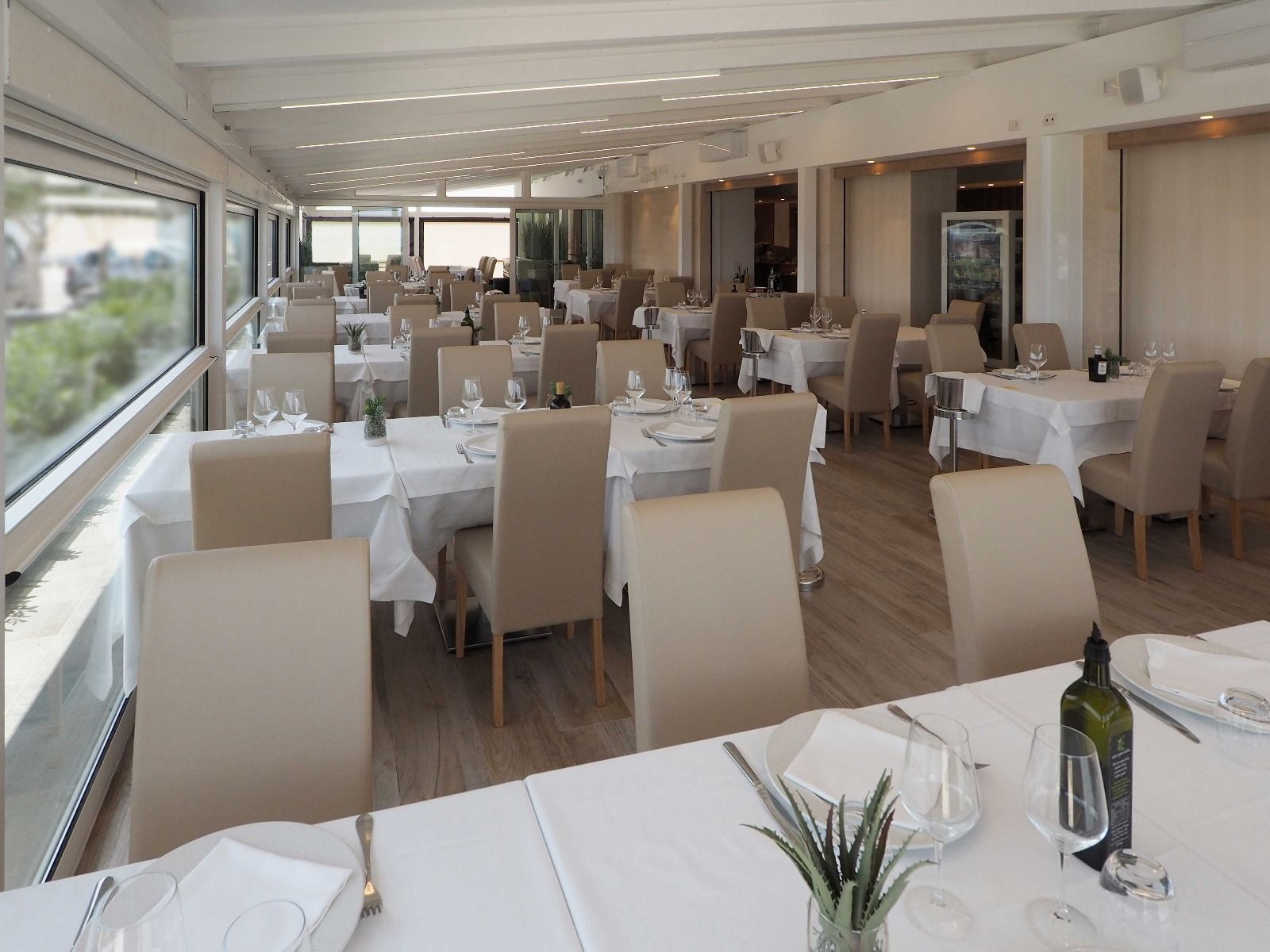 Luminosa sala da pranzo del ristorante con tovaglie bianche, sedie beige e ampie finestre che si affacciano sul verde.