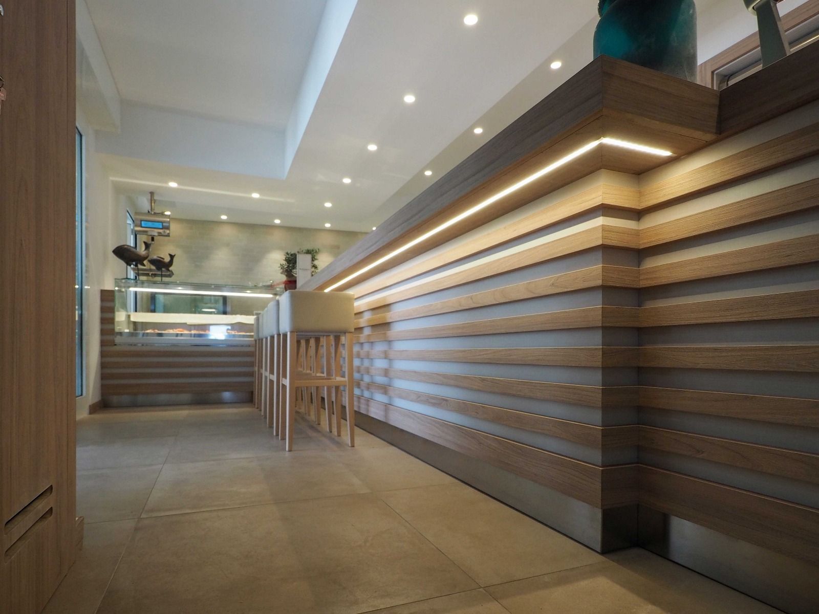 Interni moderni di un ristorante con una lunga parete in legno e metallo, illuminazione a incasso e sgabelli alti lungo un bancone.