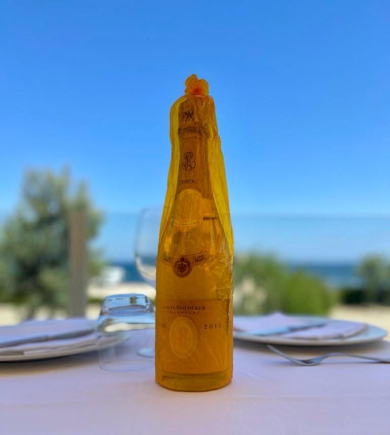 Una bottiglia color ambra su un tavolo con dei piatti, sullo sfondo di un oceano e un cielo blu sfocati.