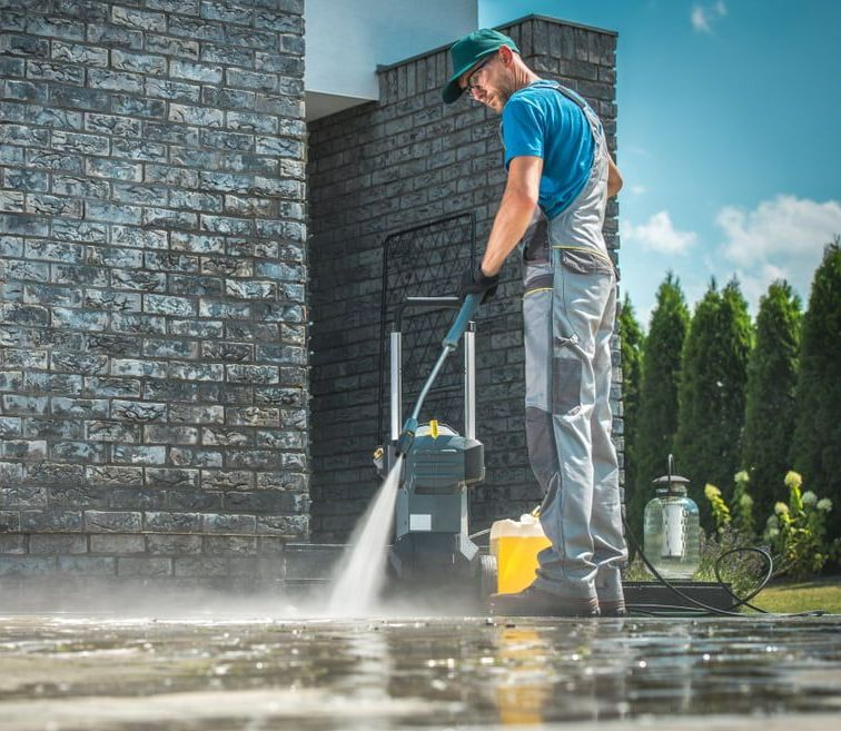 Man is Using a High Pressure Washer to Clean the Sidewalk in Front of a Brick Building — Happy Housekeepers in South Tamworth, NSW