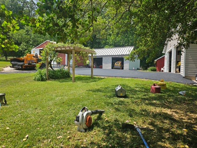 Sunny yard with a gazebo, lawn tools, a white garage, and trees in the background