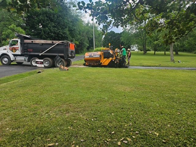 Workers paving a path beside a black dump truck and orange roller in a grassy park