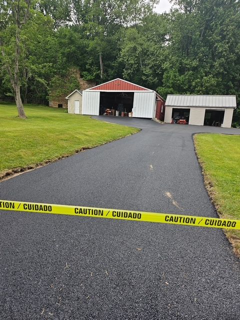 Caution tape blocks a wet driveway leading to open garage bays and a small shed in a wooded yard.