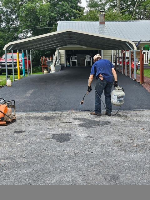 Worker spraying blacktop driveway near a carport, with a bucket and hose in hand.