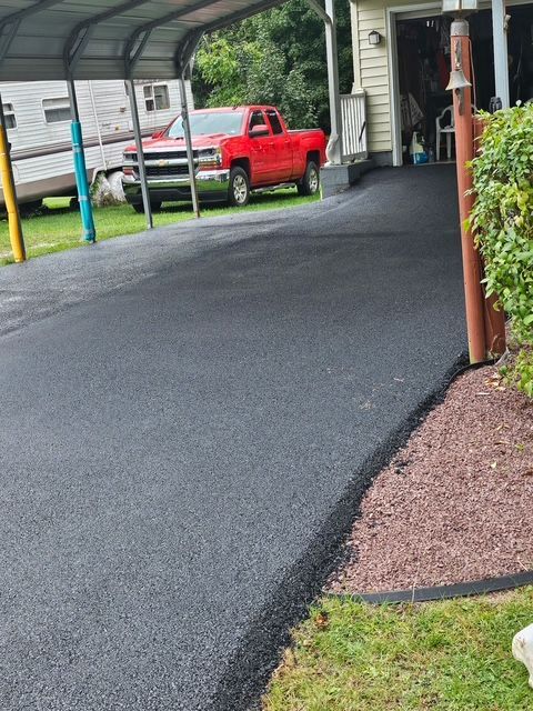 Fresh black asphalt driveway under a carport beside a red pickup truck and house.