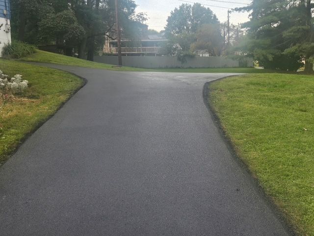 Curving paved driveway through a grassy residential area with trees and houses in the background