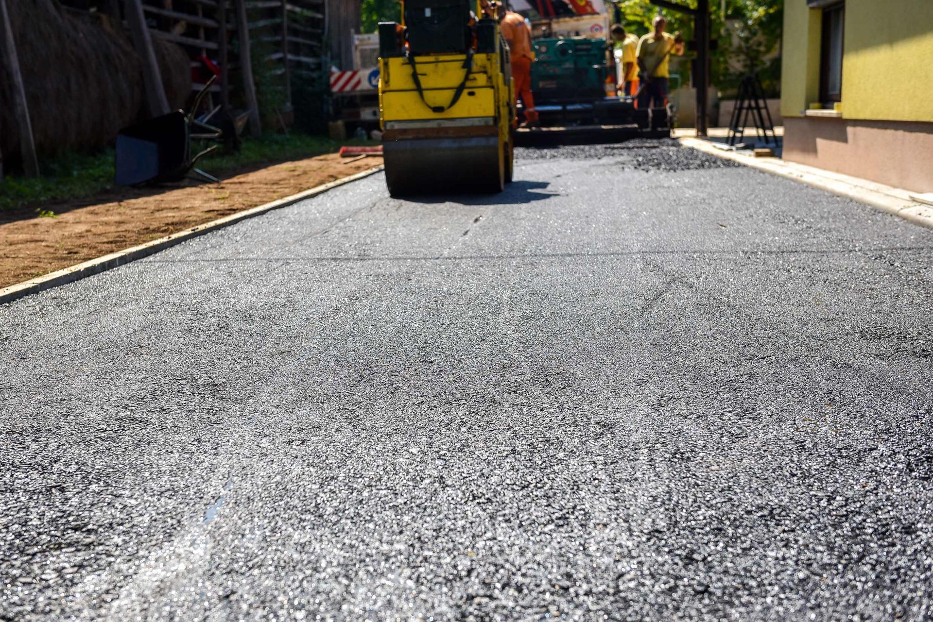 A yellow road roller compacts a fresh layer of grey asphalt on a street near a building.