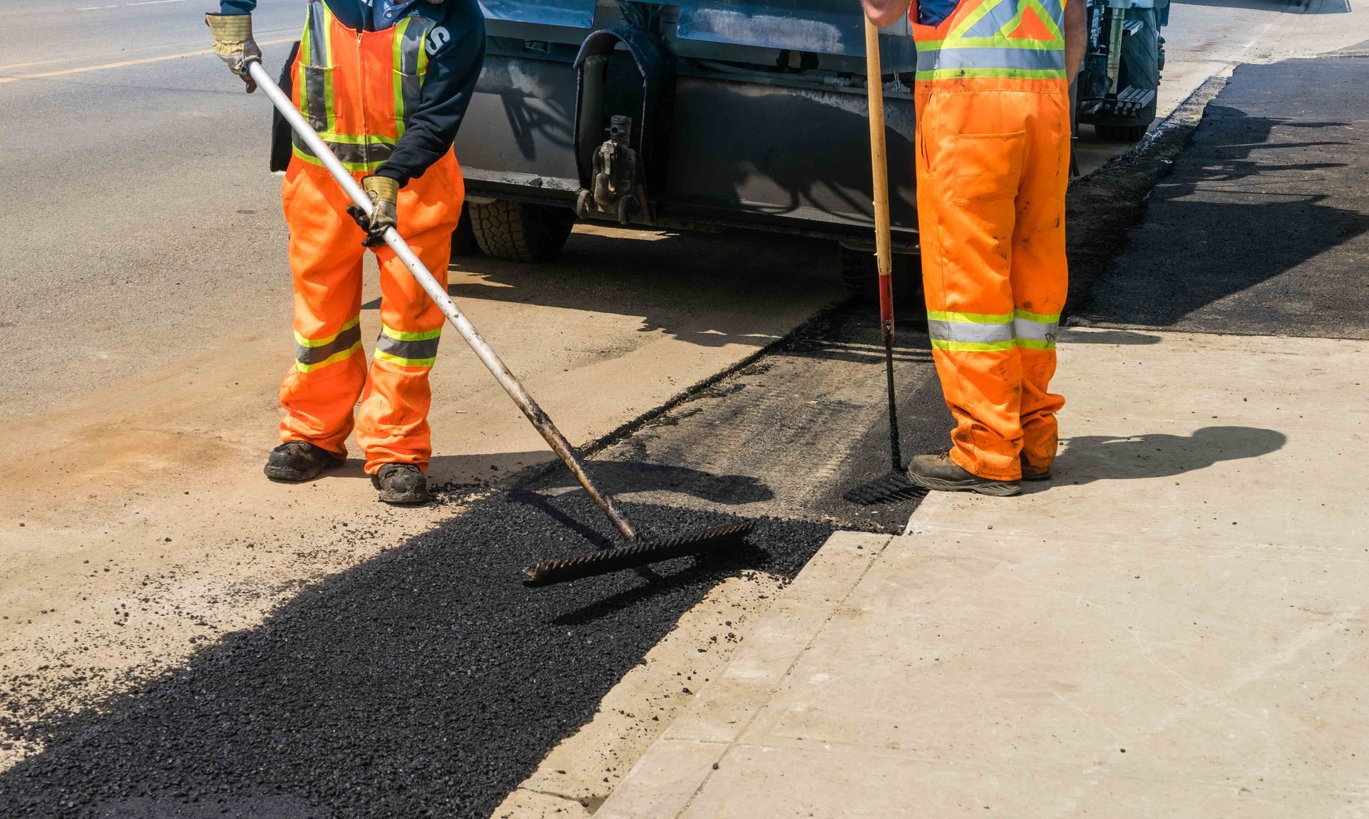 Two workers in high-visibility orange uniforms smooth fresh black asphalt into a road patch. Two workers in high-visibility orange uniforms smooth fresh black asphalt into a road patch.