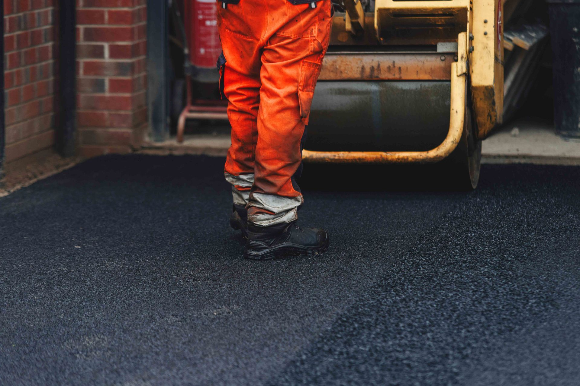 A construction worker in orange work pants stands on a freshly laid asphalt driveway next to a yellow road roller.