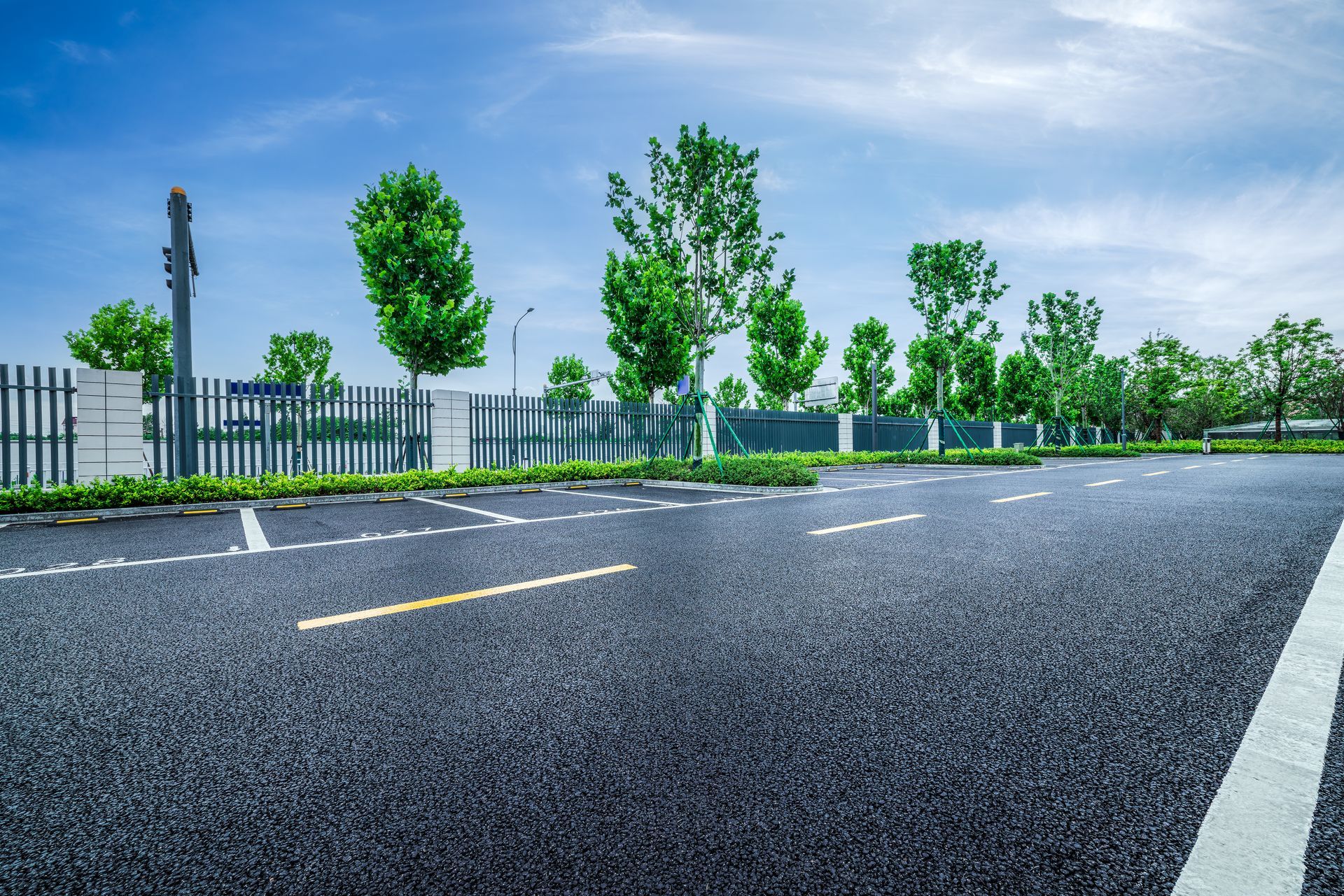 An empty parking lot with white marked spaces, lined by green trees and a fence against a clear blue sky.