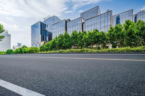 A modern glass skyscraper rises behind a row of lush green trees lining an asphalt road under a blue sky.