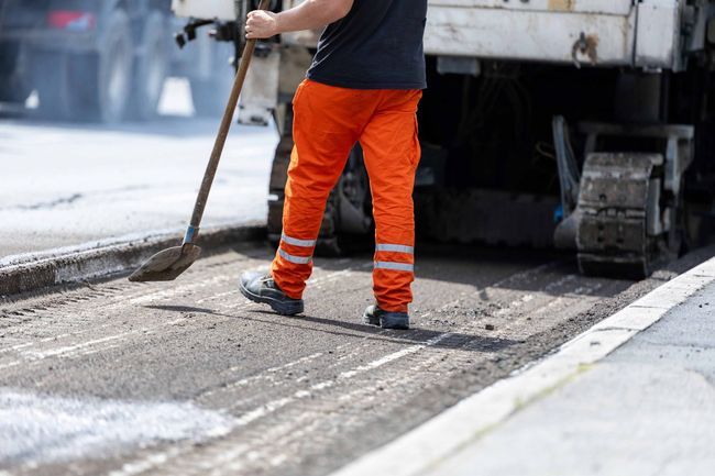 A construction worker in high-visibility orange pants uses a shovel to smooth fresh asphalt near heavy machinery.