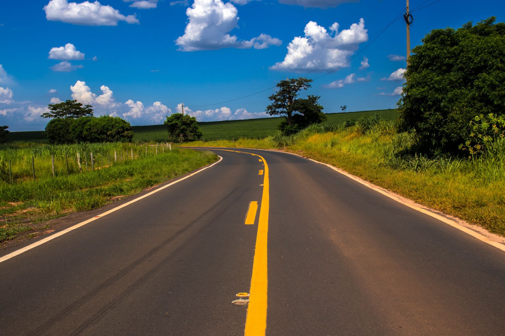 A paved road with a yellow center line curves through a lush green landscape under a bright blue, cloudy sky.