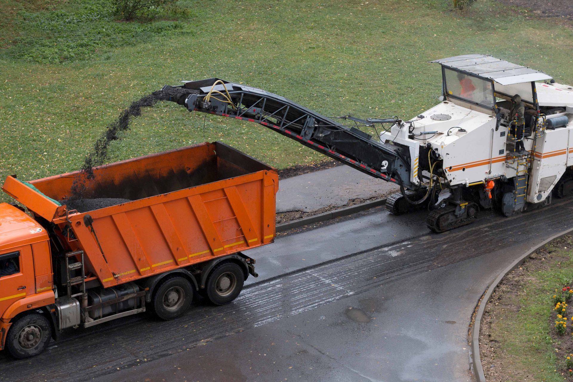 A road milling machine removes asphalt from a street, depositing the debris into the back of an orange dump truck.