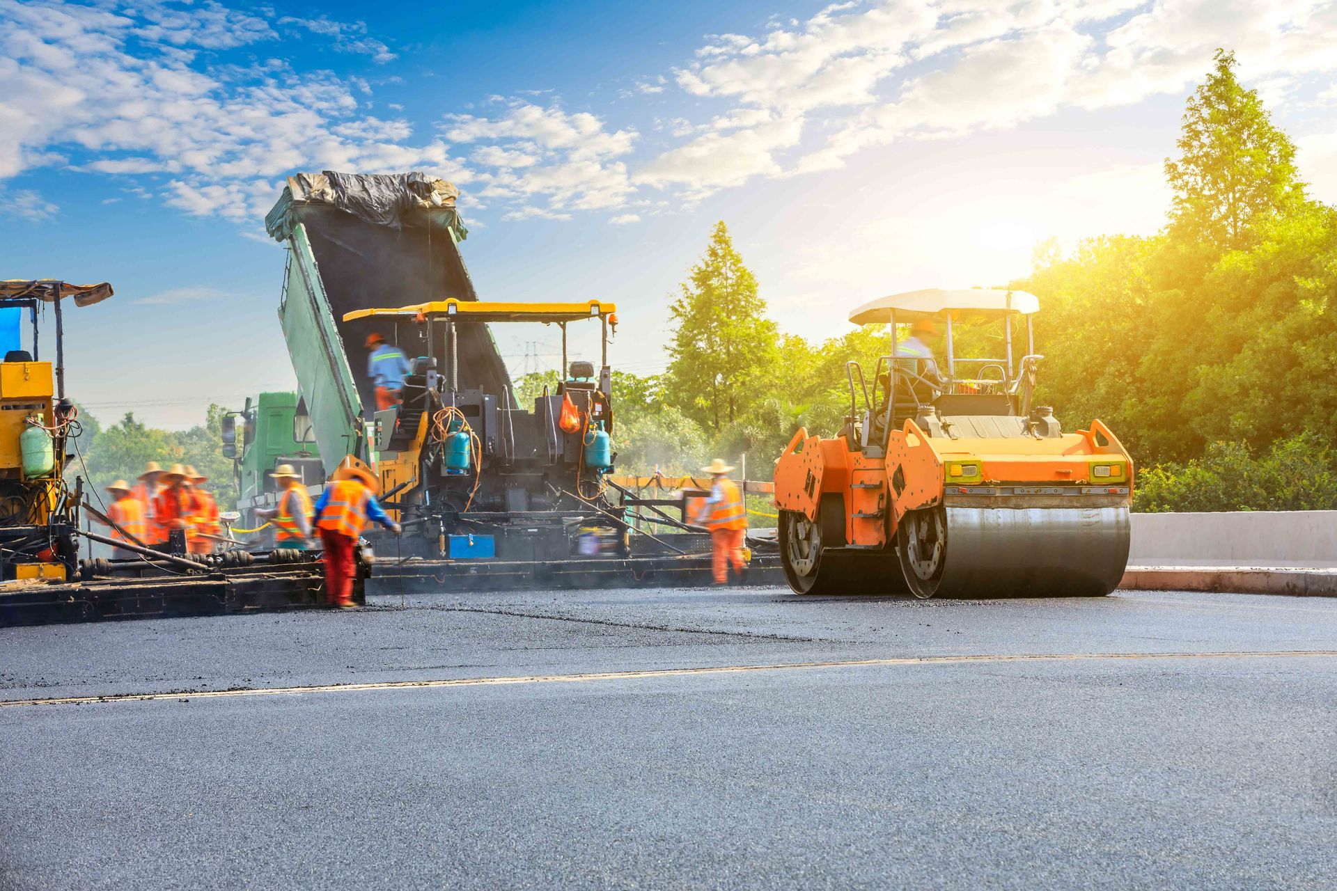 Workers pave a road with heavy machinery under a sunny sky, with a dump truck and a steamroller in operation. Workers pave a road with heavy machinery under a sunny sky, with a dump truck and a steamroller in operation.