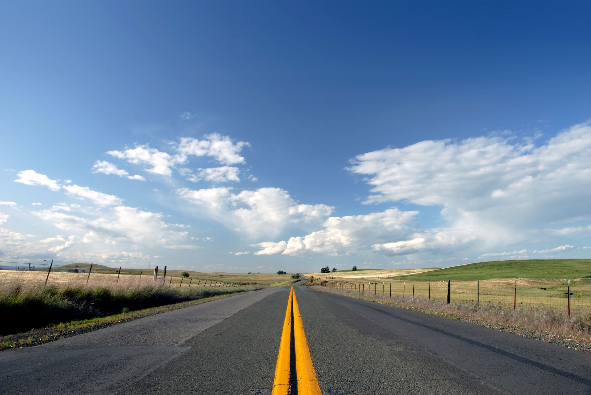 A paved rural road stretches into the distance under a bright blue sky with scattered white clouds.