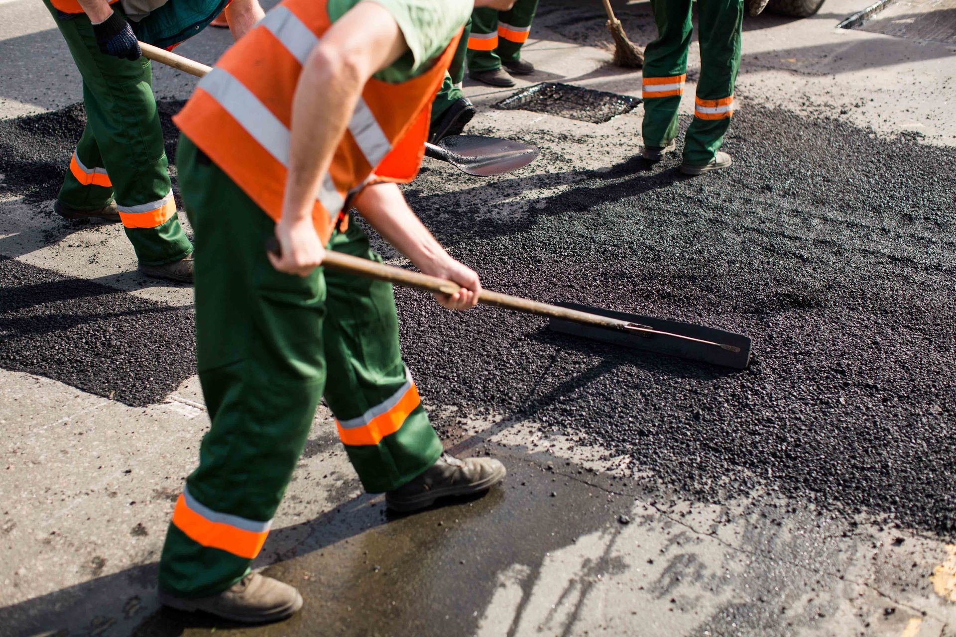Workers in orange reflective vests and green trousers use rakes to spread fresh asphalt on a road.