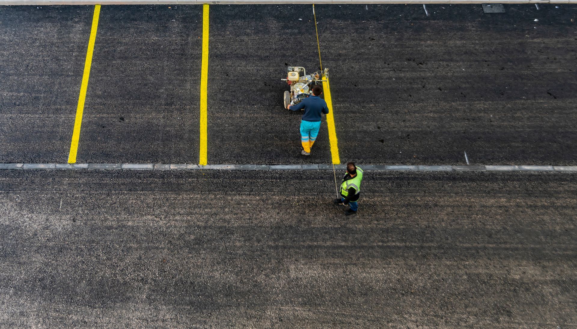 Two workers use a machine to paint yellow parking space lines on a dark asphalt lot.