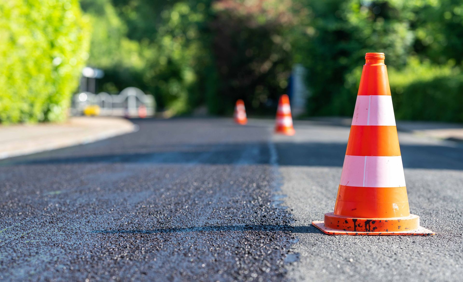 Orange and white traffic cones stand on a freshly paved asphalt road with green trees in the background. Orange and white traffic cones stand on a freshly paved asphalt road with green trees in the background.
