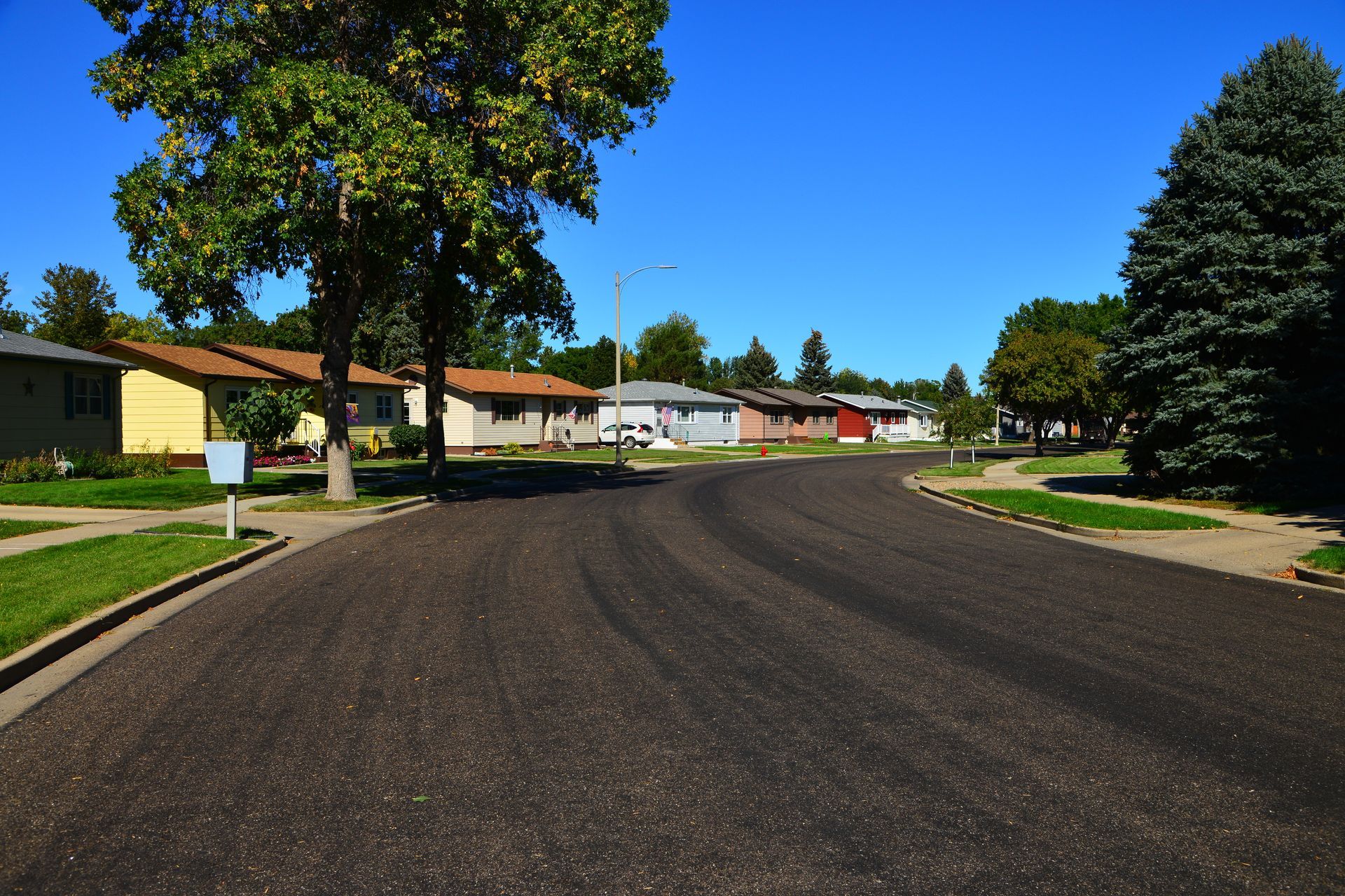 A quiet residential suburban street with a freshly paved asphalt road, green lawns, and trees under a clear blue sky.