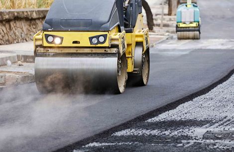 A yellow construction roller compacts fresh, dark asphalt on a road, with steam rising from the surface.
