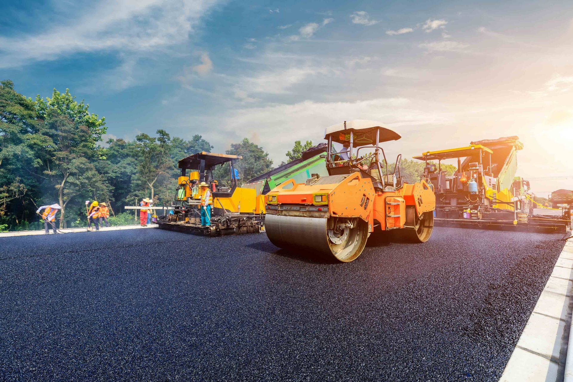 Construction workers operate yellow and orange road rollers to pave a new asphalt road at sunset. Construction workers operate yellow and orange road rollers to pave a new asphalt road at sunset.