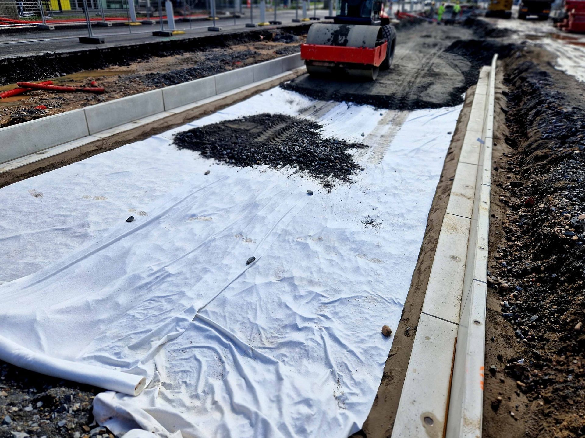 Construction workers use a roller machine to compact gravel over a white protective fabric laid in a road excavation.