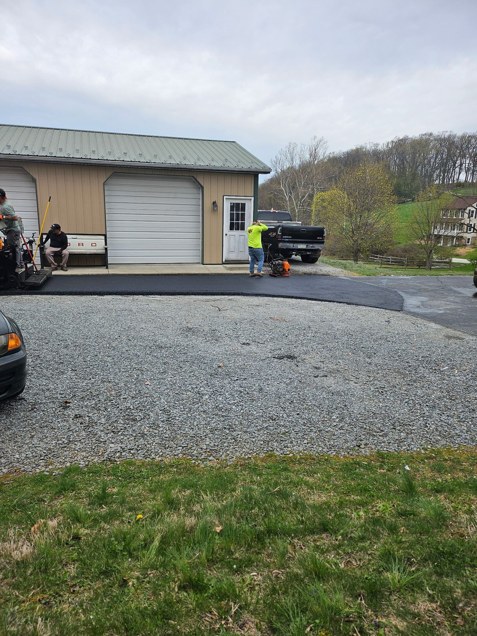 A person in work clothes spreads fresh asphalt next to a house while a yellow steamroller compacts it nearby. A person in work clothes spreads fresh asphalt next to a house while a yellow steamroller compacts it nearby.