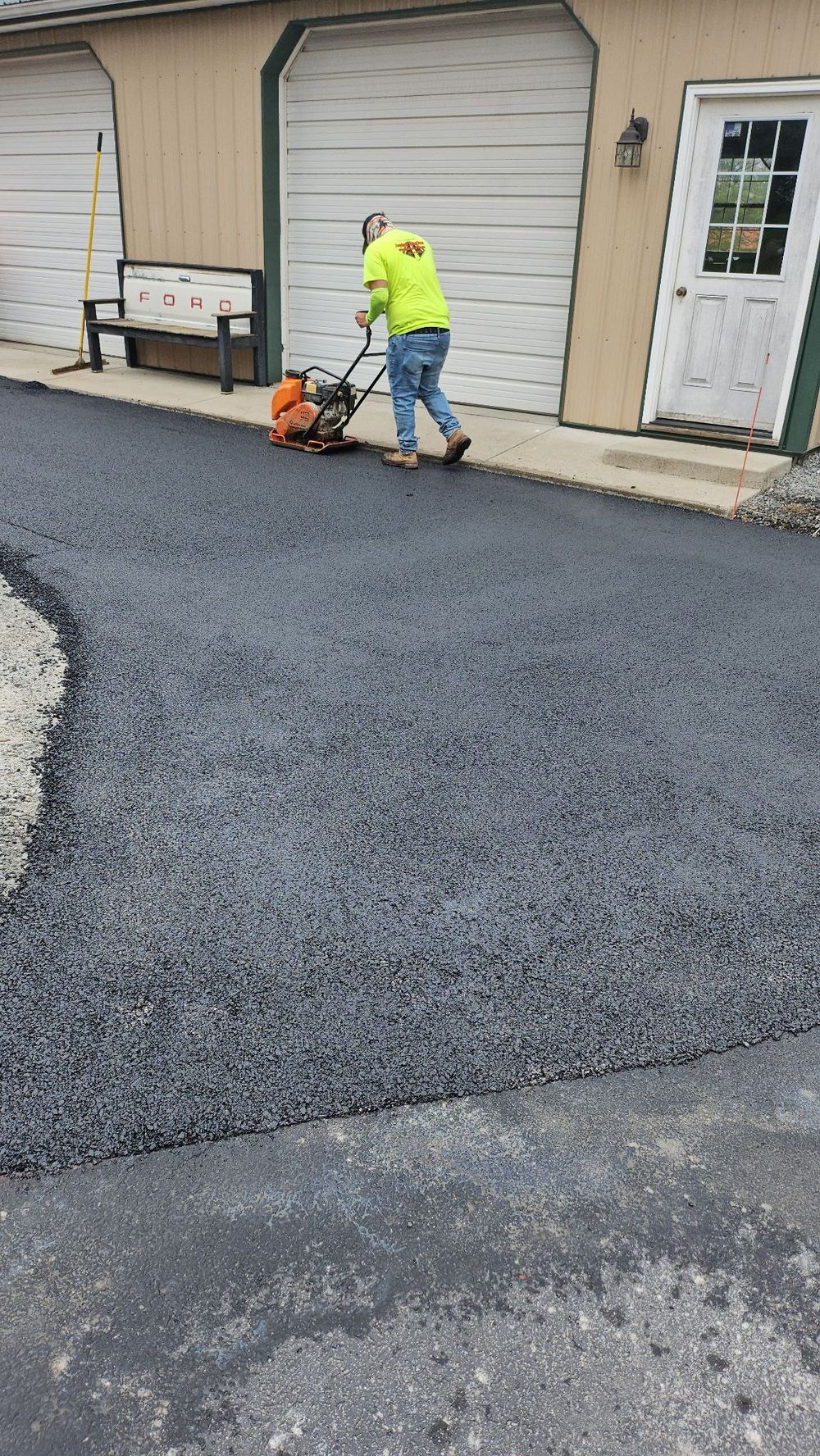 A worker in an orange safety vest smooths freshly laid black asphalt on a road near a steamroller.