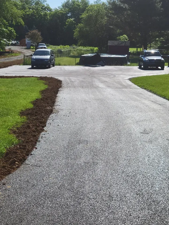 A worker in high-visibility orange pants uses a rake to spread fresh asphalt on a road surface.