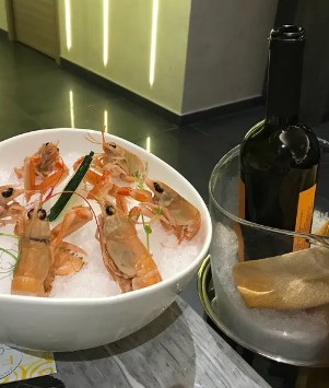 Un piatto di gamberi in una ciotola bianca accanto a una bottiglia di vino e del pane su un tavolo di ristorante.