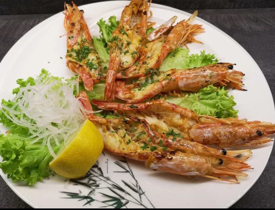 Gamberi grigliati disposti su un piatto bianco con guarnizione di lattuga, cipolla tritata e uno spicchio di limone.