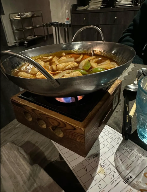Un grande wok con del cibo che sobbolle su un fornello portatile sul tavolo di un ristorante, con pinze e menù nelle vicinanze.
