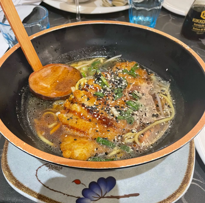 Zuppa di noodle bollente in una ciotola nera con semi di sesamo ed erbe aromatiche, servita con un cucchiaio di legno su un tavolo.