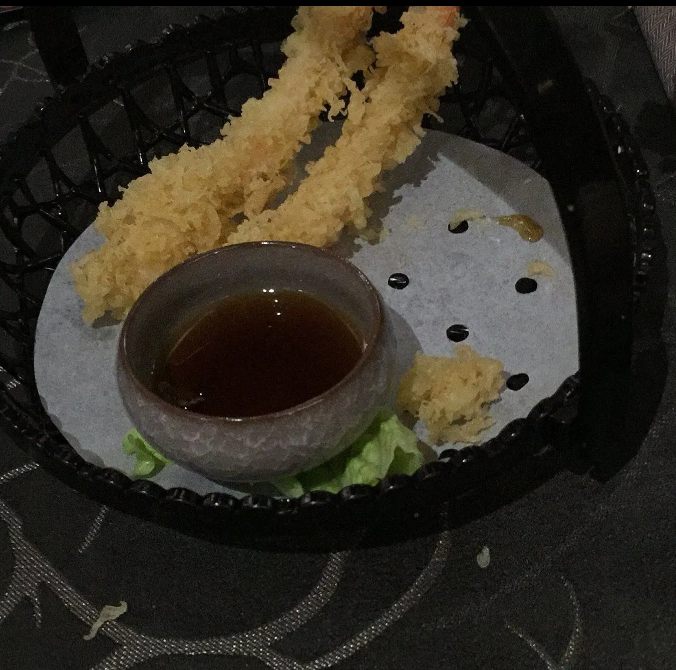 Gamberi in tempura con salsa d'accompagnamento in un contenitore nero da asporto, su un tovagliolo bianco con briciole sparse.