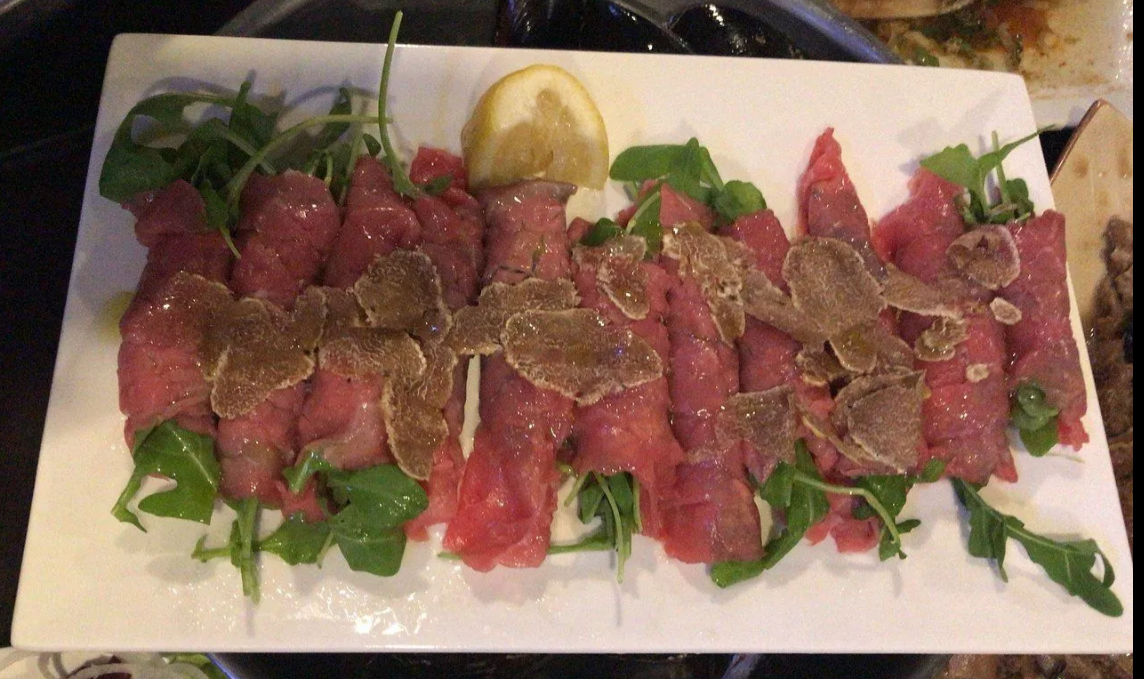 Carpaccio di manzo impiattato con rucola, scaglie di formaggio, spicchio di limone e fette di pomodoro.